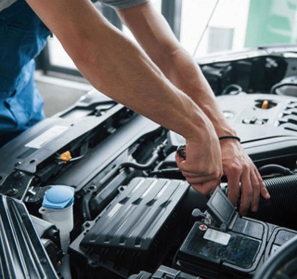 A person wearing blue overalls uses a wrench to work on a car engine under the hood, performing maintenance or repairs in a well-lit area.