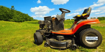 A red riding lawn mower sits on a grassy field under a blue sky with scattered clouds, bordered by trees and open greenery in the background.