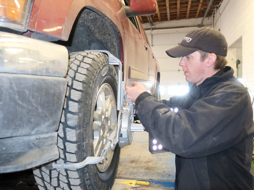 A mechanic in a black jacket and cap adjusts a wheel alignment tool attached to the front tire of a raised red SUV inside an auto repair shop.