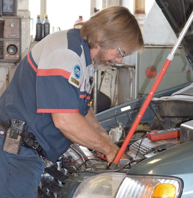 A mechanic with shoulder-length hair and glasses works on a car engine in a garage, wearing a blue uniform with patches and a radio clipped to his belt. The cars hood is open and tools are visible.