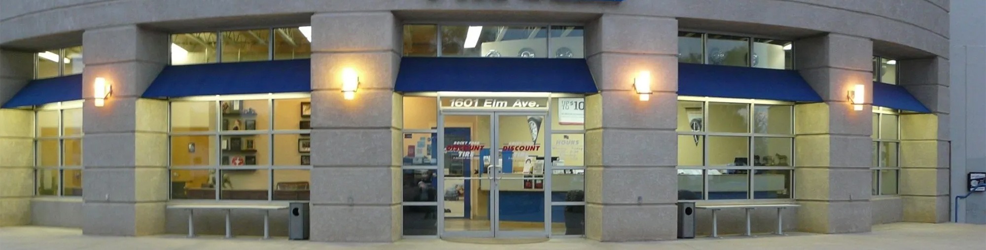 The image shows the front entrance of a modern building with large glass windows, blue awnings, two wall lights, and double glass doors. The address 1801 Film Ave. is visible above the entrance.