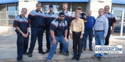 A group of ten people, some in matching work uniforms and others in casual attire, stand and pose for a photo outside a building with large windows. A sign reads Rocky Ford Discount Tire & Auto Care Center.