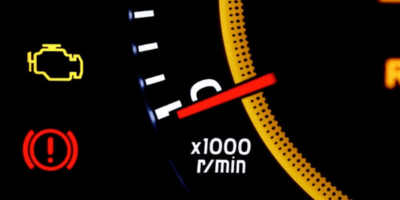 Close-up of a car dashboard showing a check engine light, a brake warning light, and a tachometer with the needle pointing near 2,000 rpm. The image is mostly dark with illuminated symbols and numbers.