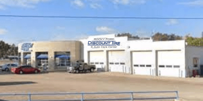 A discount tire and auto care center with a main entrance, multiple garage bays, parked vehicles, and a clear blue sky in the background.