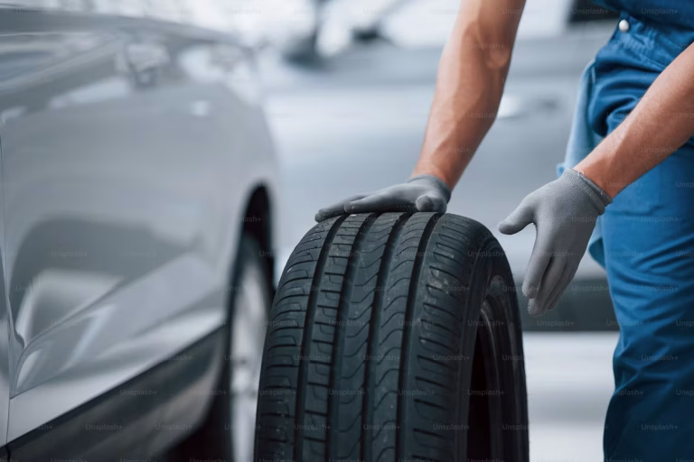 A person wearing gray gloves rolls a car tire next to a silver vehicle, preparing for installation or replacement in an automotive service setting.
