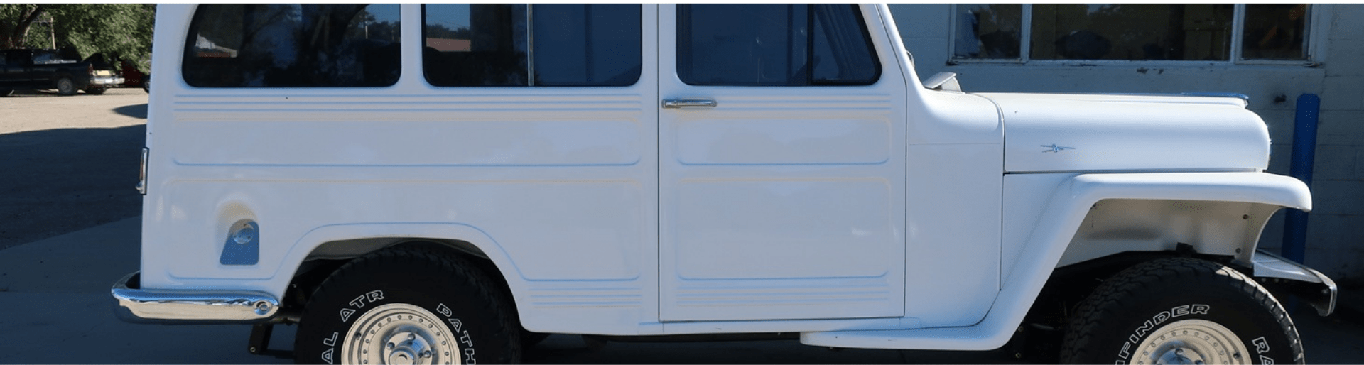 A classic white vintage SUV with large off-road tires is parked beside a building, partially in the shade on a sunny day.