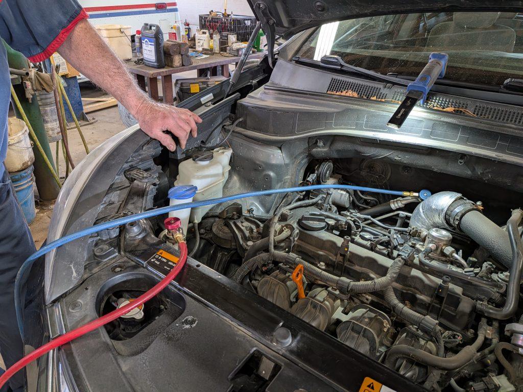 A person services a car engine in a workshop, with the hood open. Hoses are connected to the battery and fluid reservoir, and tools and equipment are visible in the background.