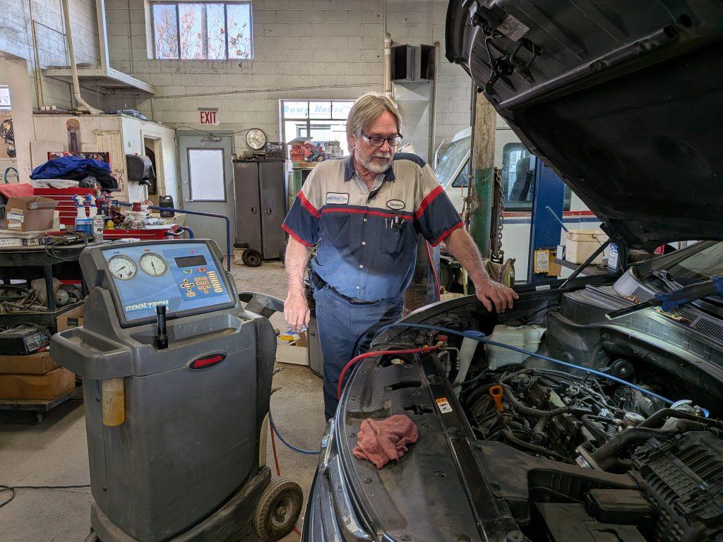 A mechanic wearing glasses and a work uniform inspects a car engine in a garage. Diagnostic equipment is positioned next to the car, and various tools and supplies are visible in the background.