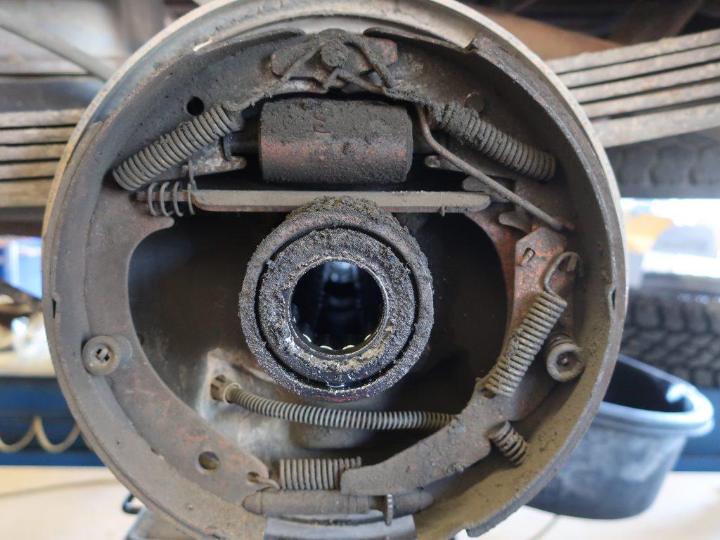 Close-up of a dusty and worn drum brake assembly on a vehicle, showing springs, brake shoes, and a central circular hub, with visible rust and grime buildup.