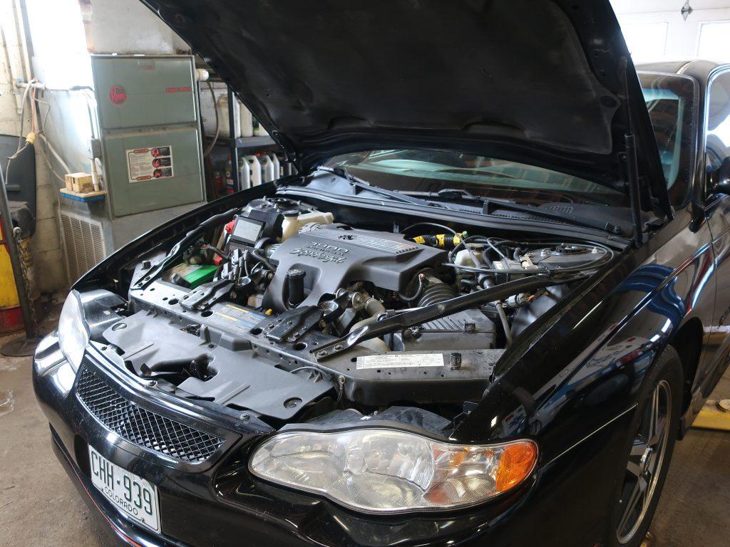 A black car with its hood open reveals the engine and various components inside a garage. Tools, equipment, and containers are visible in the background. The car’s front license plate reads “CHH-939.”.