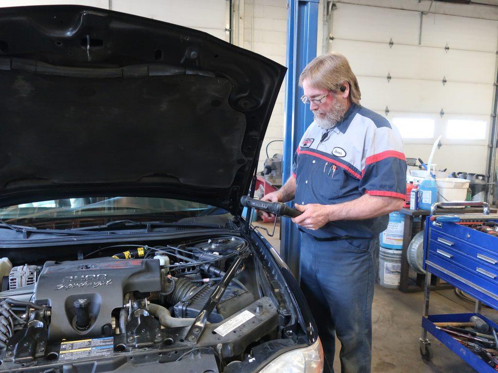 An auto mechanic wearing a work uniform inspects a car engine with the hood open, using a diagnostic tool in a garage with tools and equipment in the background.