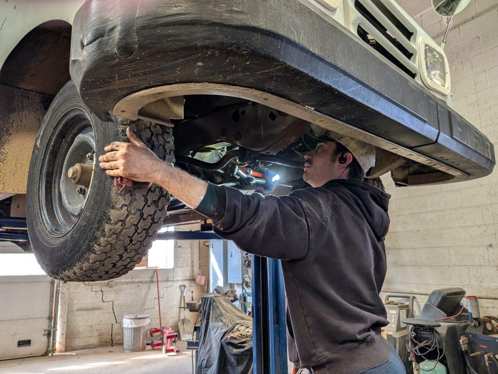 A mechanic wearing a hoodie and cap inspects and works on the front wheel area of a raised vehicle in a garage workshop, focusing on suspension and steering repair. Tools and equipment are visible in the background.