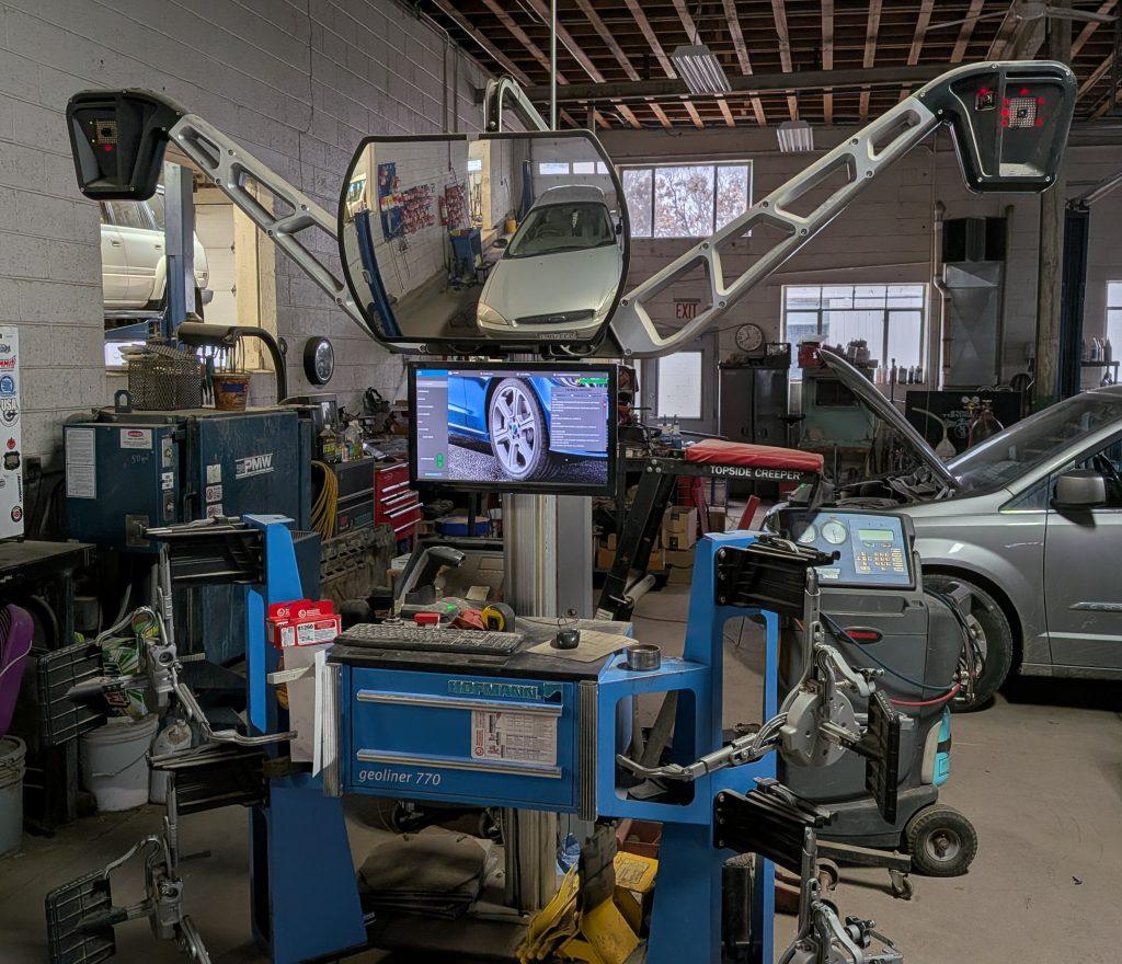A wheel alignment machine with monitors and sensors is set up in an auto repair shop for precise wheel alignments, with cars parked inside and various tools and equipment visible in the background.