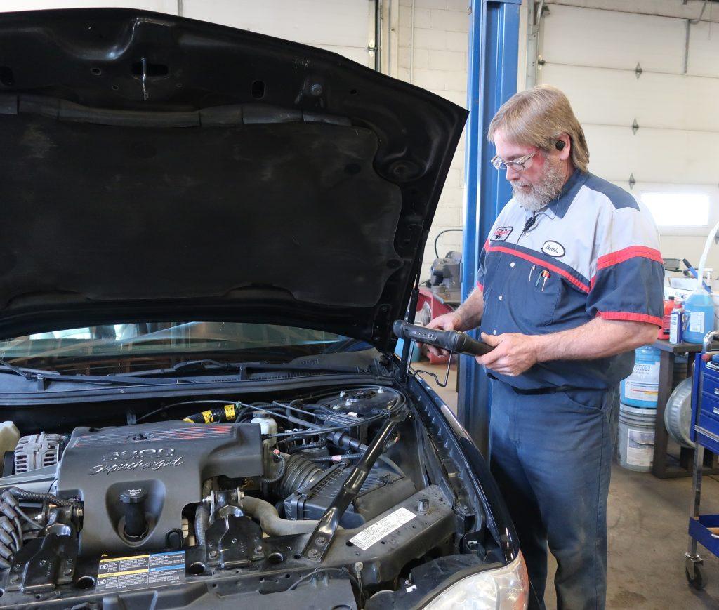 A mechanic with glasses and a beard inspects a car engine with the hood open, using a handheld computer diagnostics tool in an auto repair shop.