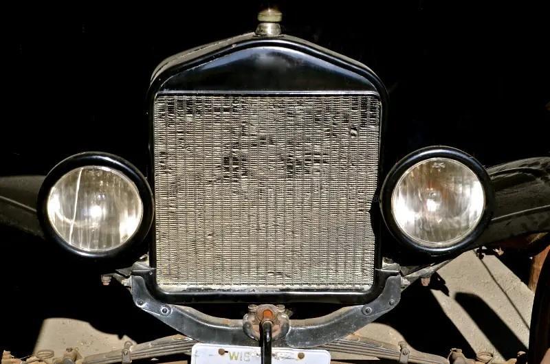 Close-up of the front of a vintage car showing a shiny metal grille, two round headlights, and part of a license plate. The background is dark, highlighting the classic features and hinting at expert cooling system service and repair.
