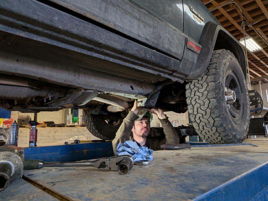 A mechanic wearing a cap works underneath a raised SUV in a garage, using a tool on the vehicle’s undercarriage—perfect for tasks like wheel alignments. Various tools and a cloth rest on the platform nearby.