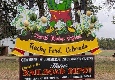A colorful sign reads Home of the Meloneers, Sweet Melon Capital, Rocky Ford, Colorado. It features a flexing watermelon cartoon and info about the Chamber of Commerce at the historic Railroad Depot.