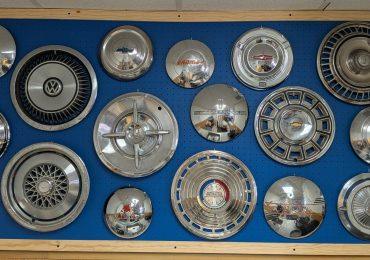 A blue pegboard displays a collection of vintage car hubcaps, including brands like Volkswagen, Chevrolet, Nissan, and Plymouth, mounted neatly in rows within a wooden frame.